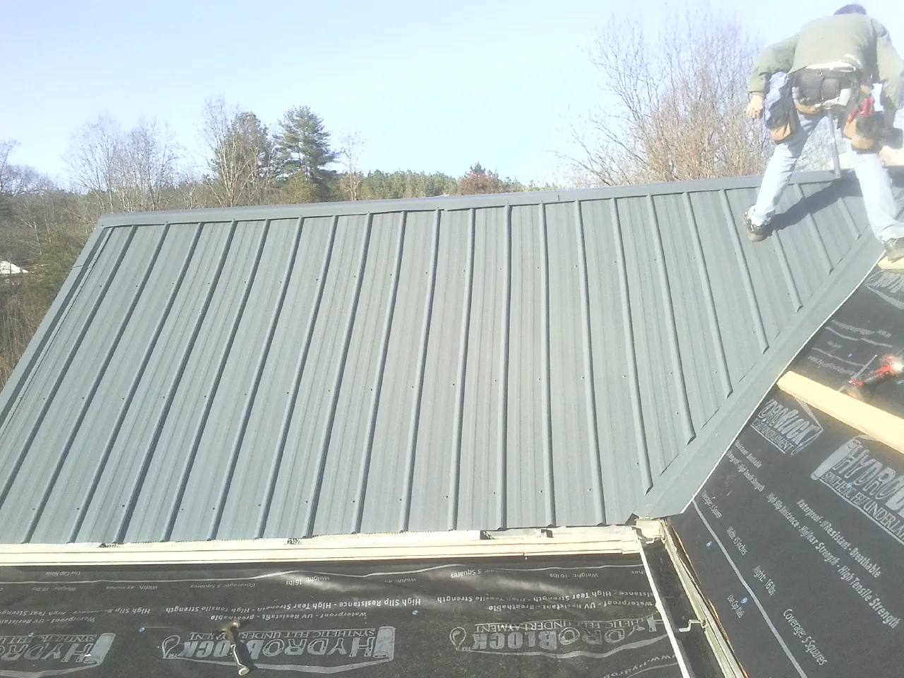 Crew installing dark green standing seam panels over synthetic underlayment in Mason City