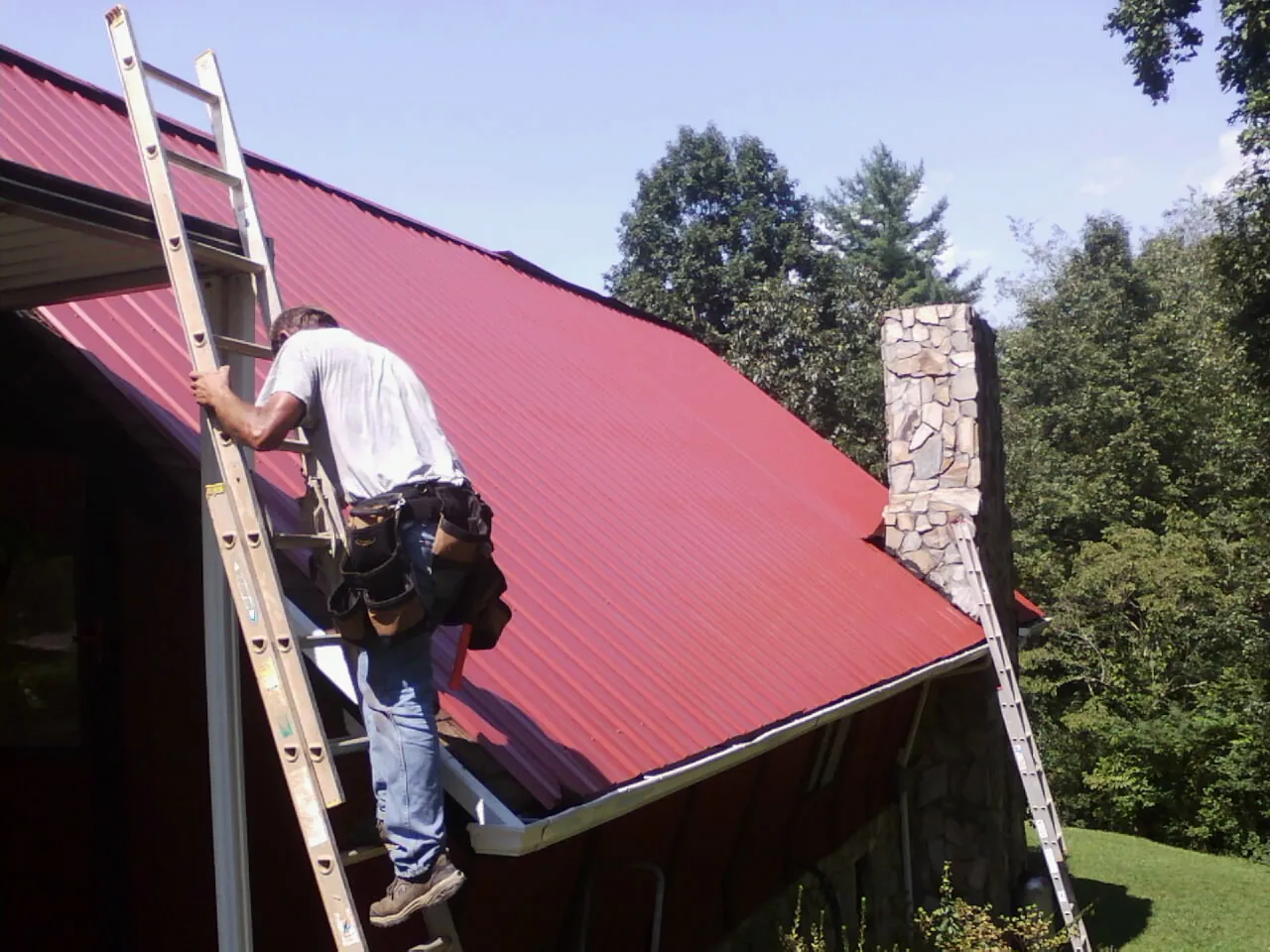 Roofer on a ladder beside a red metal roof and stone chimney — Soffit & Fascia Repair by Roofing Brothers in Mason City